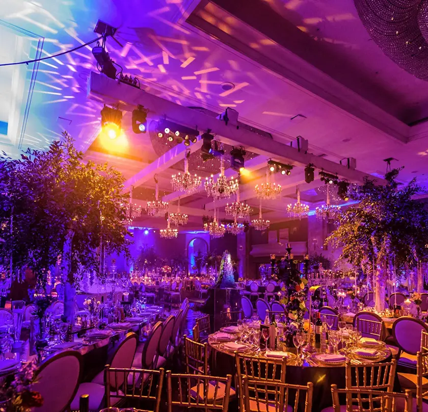 Lavish wedding reception with elegant chandeliers, colorful lighting, and beautifully set round tables.